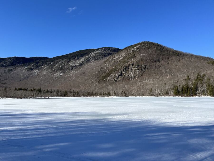 A snow-covered frozen pond below a wooded mountain